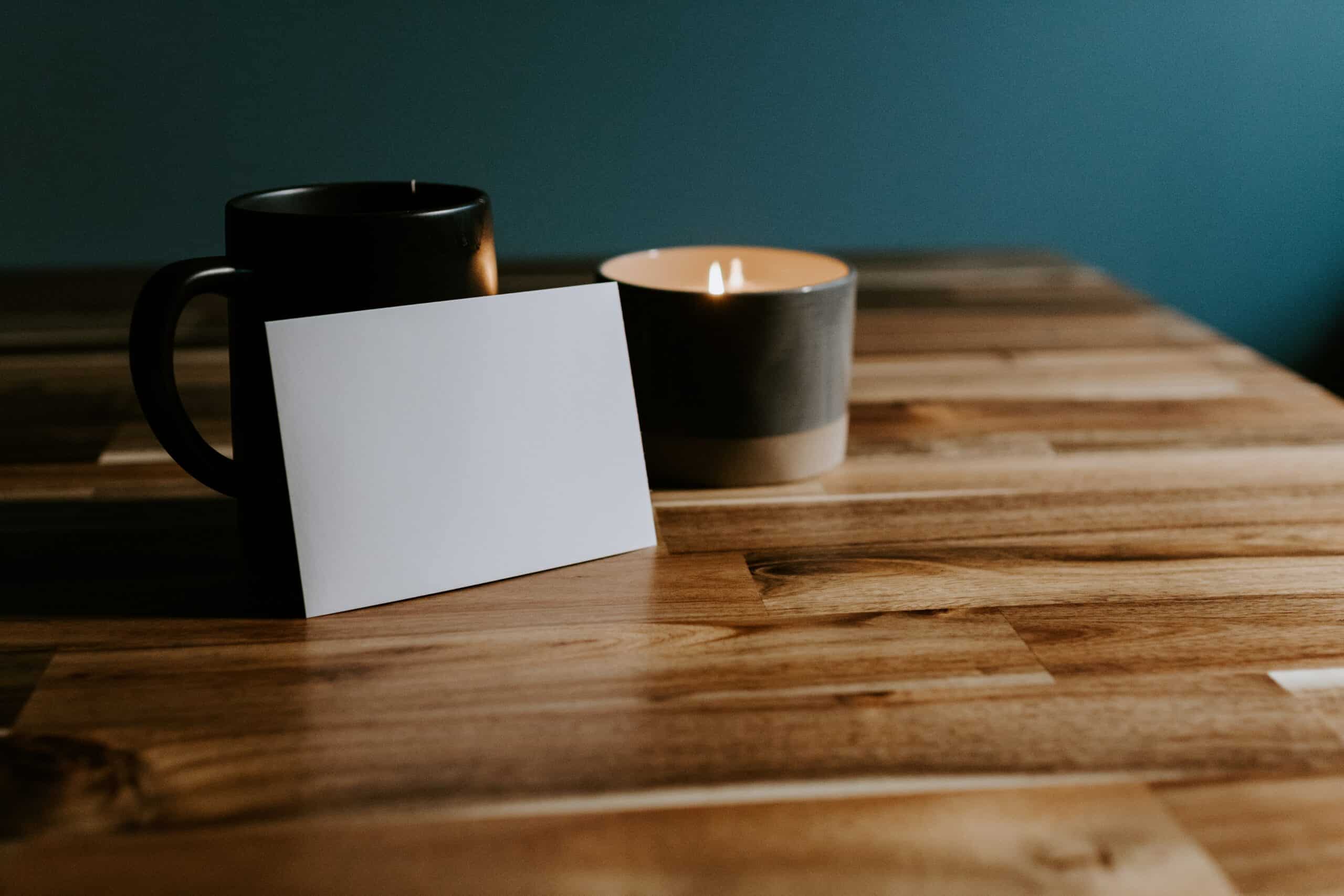 A mug, candle, and paper card, sitting on top of wooden table top